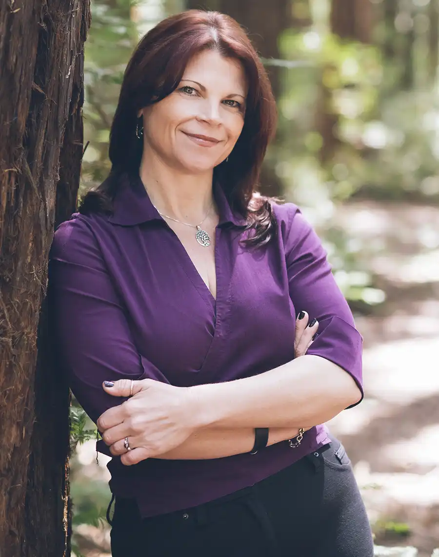 Headshot of April Bell wearing a eggplant blouse, leaning against a redwood tree in a redwood forest. Dappled sunlight in background.
